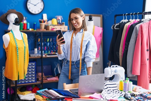 Young beautiful hispanic woman tailor using smartphone and laptop at clothing factory