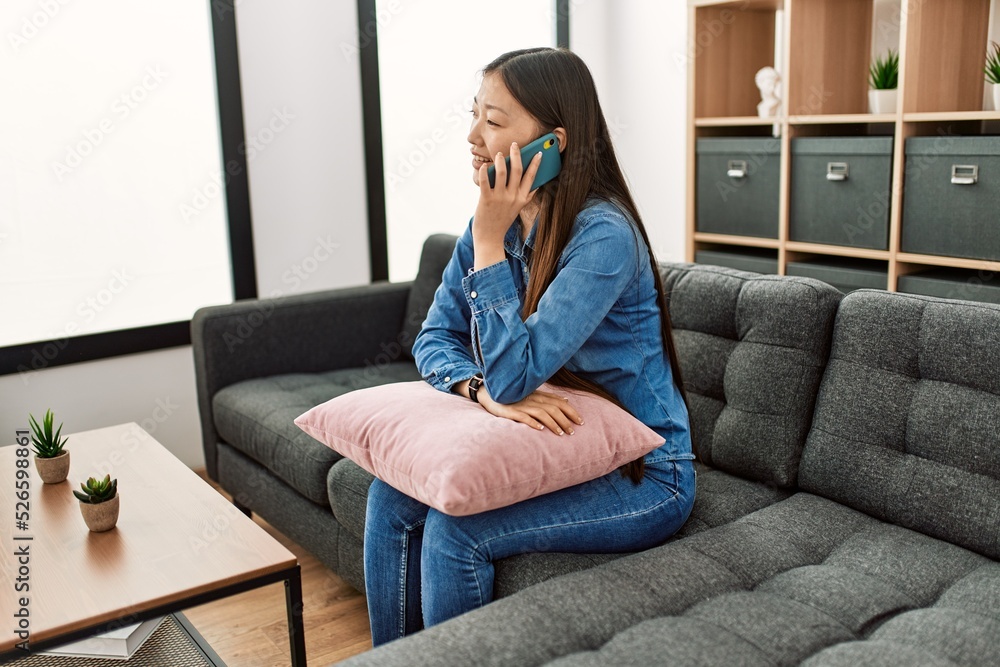 Young chinese girl talking on the smartphone sitting on the sofa at home.