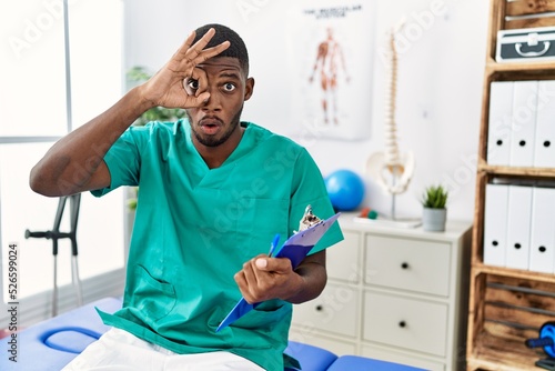 Young african american man working at pain recovery clinic doing ok gesture shocked with surprised face  eye looking through fingers. unbelieving expression.