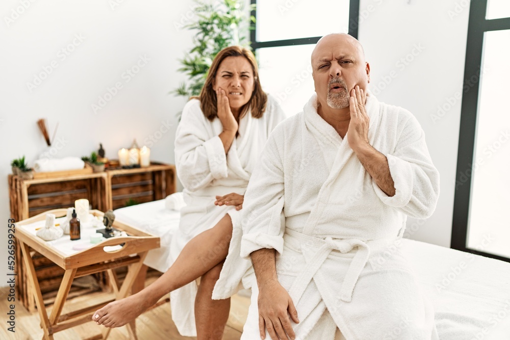 Middle age hispanic couple wearing bathrobe at wellness spa touching mouth with hand with painful expression because of toothache or dental illness on teeth. dentist Middle age hispanic couple wearing bathrobe at wellness spa touching mouth with hand with painful expression because of toothache or dental illness on teeth. dentist