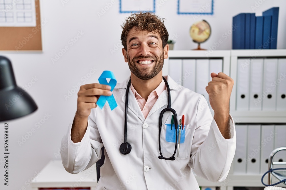 Young hispanic doctor man holding blue ribbon screaming proud, celebrating victory and success very excited with raised arms Young hispanic doctor man holding blue ribbon screaming proud, celebrating victory and success very excited with raised arms