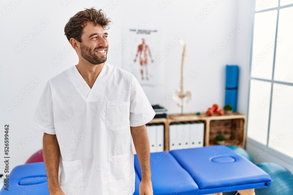 Young handsome physiotherapist man working at pain recovery clinic looking away to side with smile on face, natural expression. laughing confident. Young handsome physiotherapist man working at pain recovery clinic looking away to side with smile on face, natural expression. laughing confident.