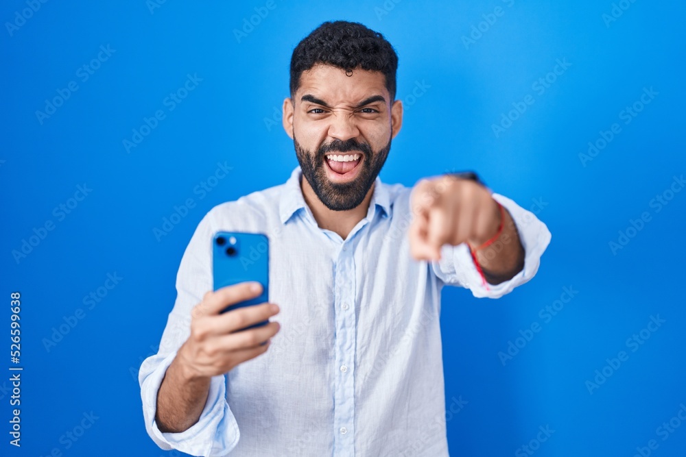 Hispanic man with beard using smartphone typing message pointing displeased and frustrated to the camera, angry and furious with you Hispanic man with beard using smartphone typing message pointing displeased and frustrated to the camera, angry and furious with you