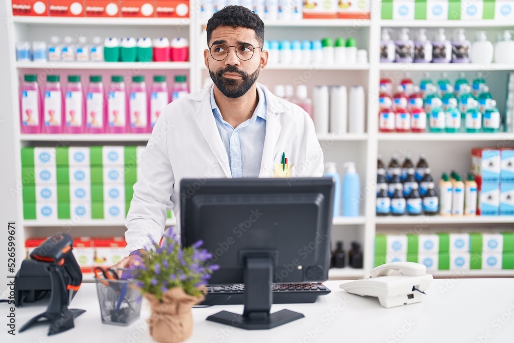 Hispanic man with beard working at pharmacy drugstore smiling looking to the side and staring away thinking.