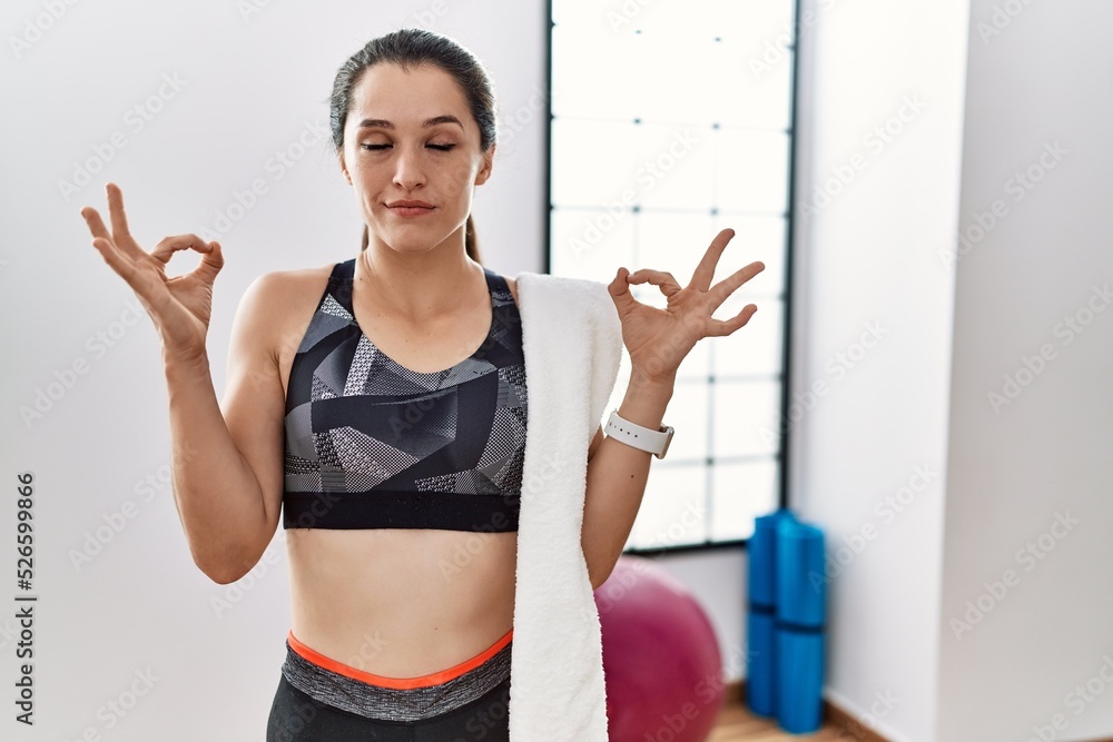 Young brunette woman wearing sportswear and towel at the gym relaxed and smiling with eyes closed doing meditation gesture with fingers. yoga concept.