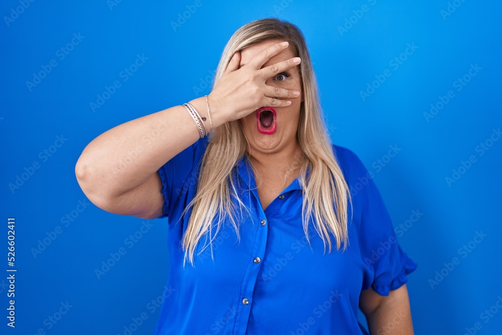 Caucasian plus size woman standing over blue background peeking in shock covering face and eyes with hand, looking through fingers with embarrassed expression. Caucasian plus size woman standing over blue background peeking in shock covering face and eyes with hand, looking through fingers with embarrassed expression.