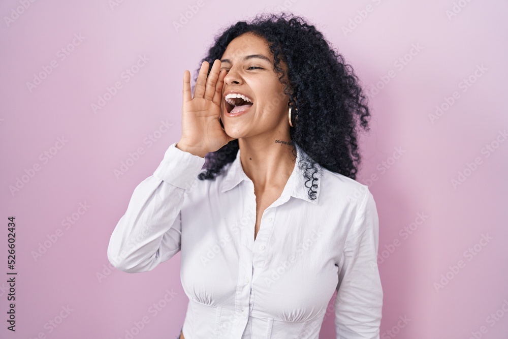 Hispanic woman with curly hair standing over pink background shouting and screaming loud to side with hand on mouth. communication concept. Hispanic woman with curly hair standing over pink background shouting and screaming loud to side with hand on mouth. communication concept.