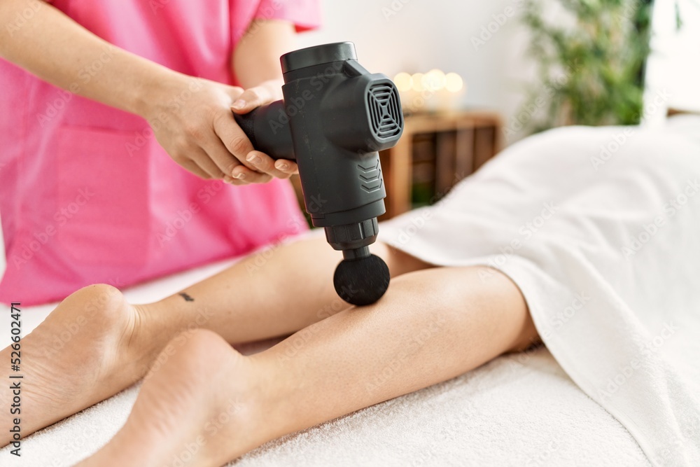 Young hispanic woman having legs massage with percussion gun at beauty center.