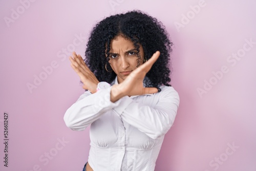 Hispanic woman with curly hair standing over pink background rejection expression crossing arms doing negative sign, angry face © Krakenimages.com