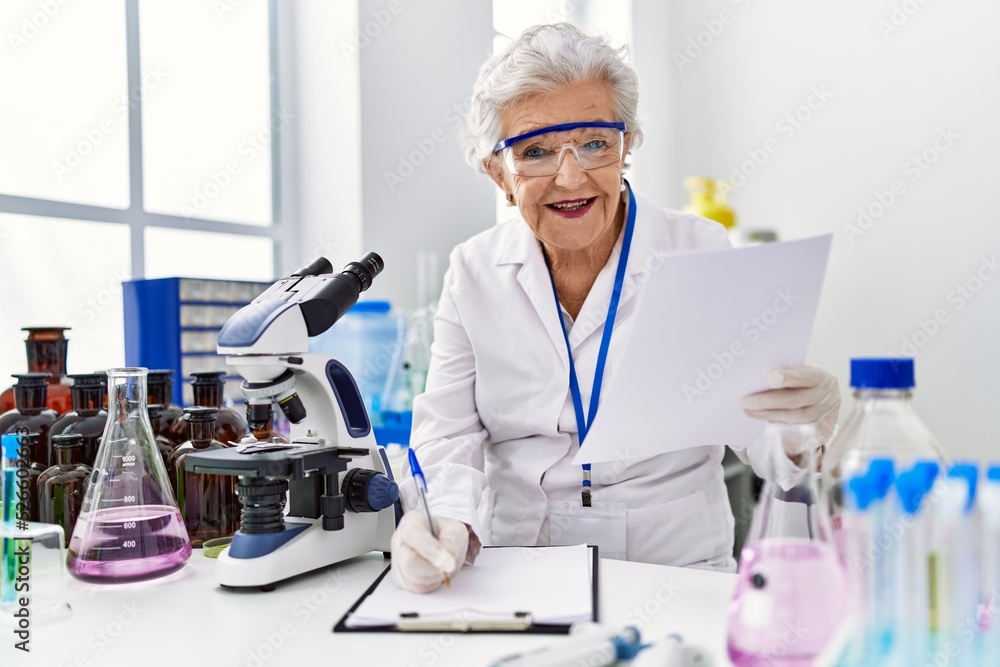 Senior grey-haired woman wearing scientist uniform writing on clipboard at laboratory Senior grey-haired woman wearing scientist uniform writing on clipboard at laboratory