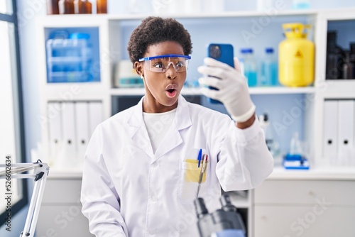 African american woman working at scientist laboratory doing video call with smartphone scared and amazed with open mouth for surprise, disbelief face