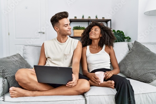 Young interracial couple using laptop at home sitting on the sofa smiling looking to the side and staring away thinking.