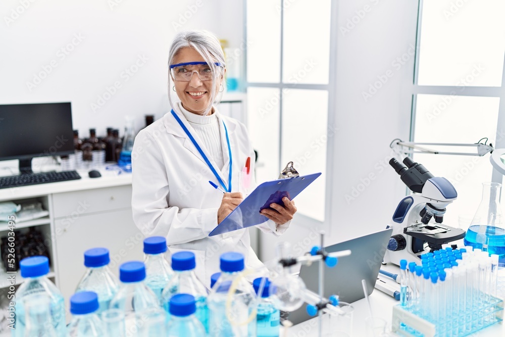 Fototapeta premium Middle age grey-haired woman wearing scientist uniform writing on clipboard working at laboratory