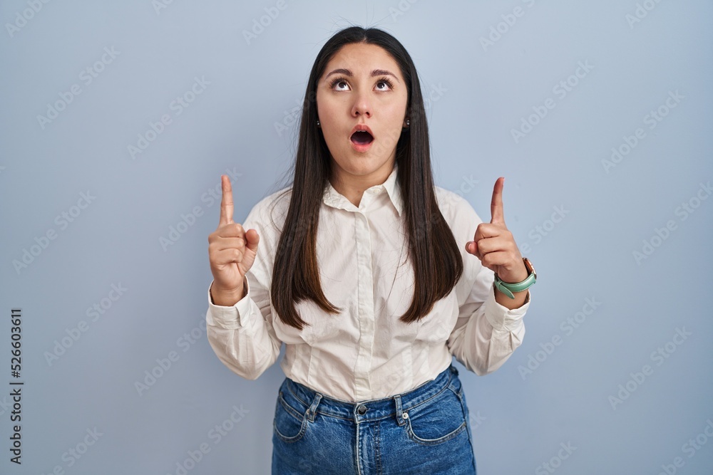 Young latin woman standing over blue background amazed and surprised looking up and pointing with fingers and raised arms. Young latin woman standing over blue background amazed and surprised looking up and pointing with fingers and raised arms.