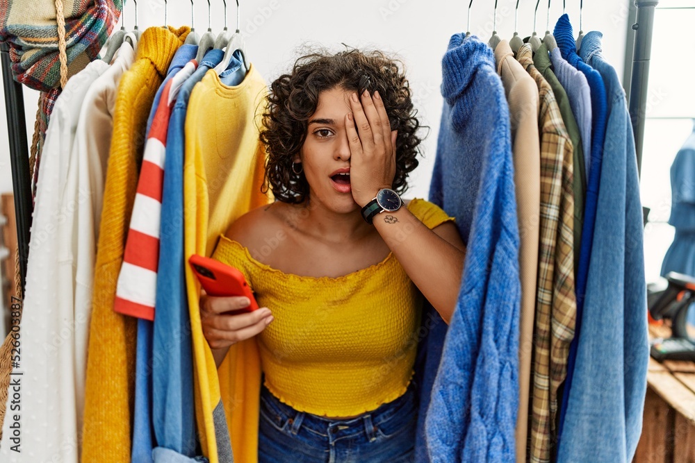 Young hispanic woman searching clothes on clothing rack using smartphone yawning tired covering half face, eye and mouth with hand. face hurts in pain. Young hispanic woman searching clothes on clothing rack using smartphone yawning tired covering half face, eye and mouth with hand. face hurts in pain.
