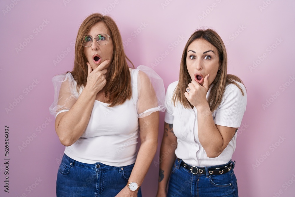 Hispanic mother and daughter wearing casual white t shirt over pink background looking fascinated with disbelief, surprise and amazed expression with hands on chin Hispanic mother and daughter wearing casual white t shirt over pink background looking fascinated with disbelief, surprise and amazed expression with hands on chin