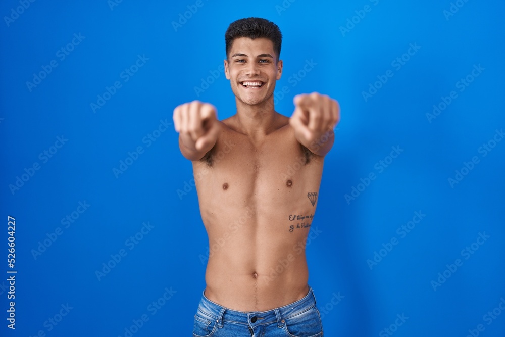 Young hispanic man standing shirtless over blue background pointing to you and the camera with fingers, smiling positive and cheerful