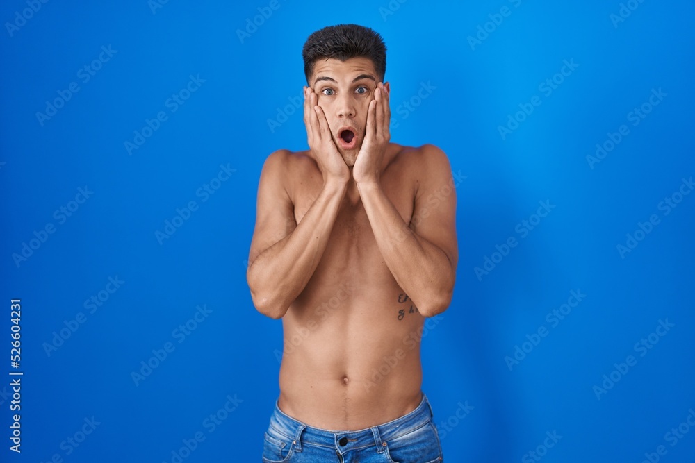 Fototapeta premium Young hispanic man standing shirtless over blue background afraid and shocked, surprise and amazed expression with hands on face