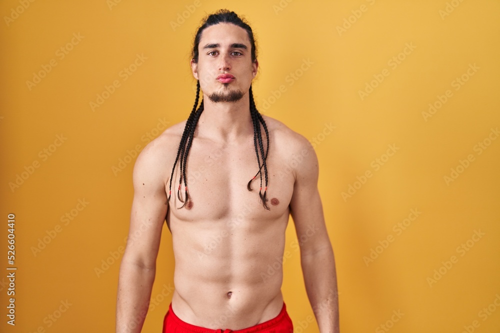 Fototapeta premium Hispanic man with long hair standing shirtless over yellow background looking at the camera blowing a kiss on air being lovely and sexy. love expression.