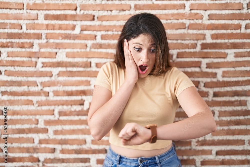 Young brunette woman standing over bricks wall looking at the watch time worried, afraid of getting late