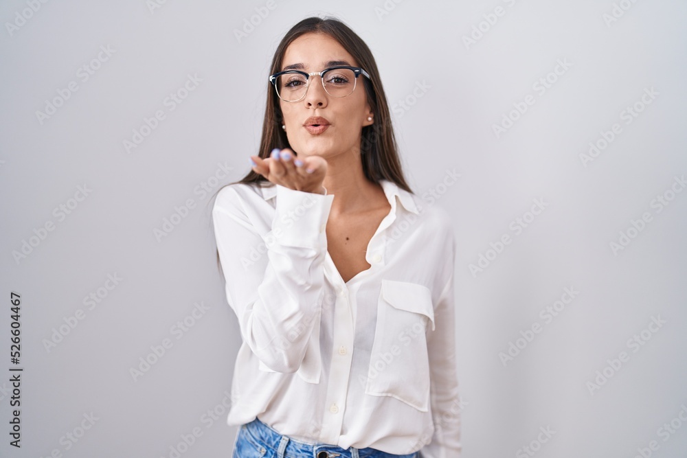 Young brunette woman wearing glasses looking at the camera blowing a kiss with hand on air being lovely and sexy. love expression. Young brunette woman wearing glasses looking at the camera blowing a kiss with hand on air being lovely and sexy. love expression.