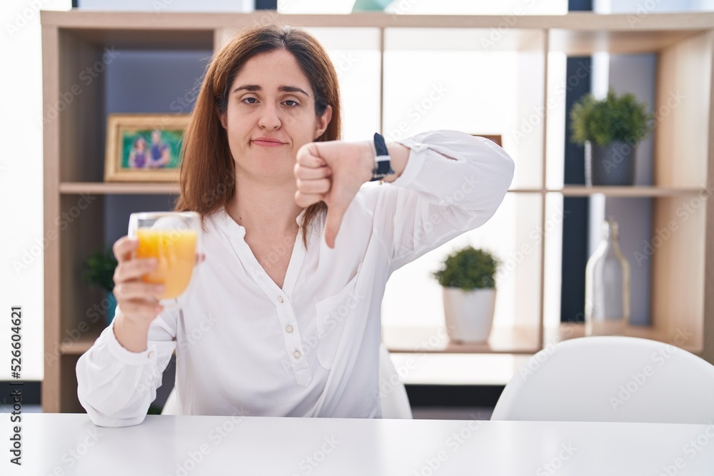 Brunette woman drinking glass of orange juice looking unhappy and angry showing rejection and negative with thumbs down gesture. bad expression. Brunette woman drinking glass of orange juice looking unhappy and angry showing rejection and negative with thumbs down gesture. bad expression.
