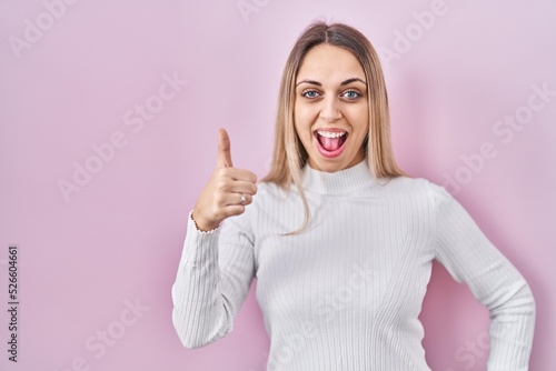Young blonde woman wearing white sweater over pink background doing happy thumbs up gesture with hand. approving expression looking at the camera showing success. © Krakenimages.com