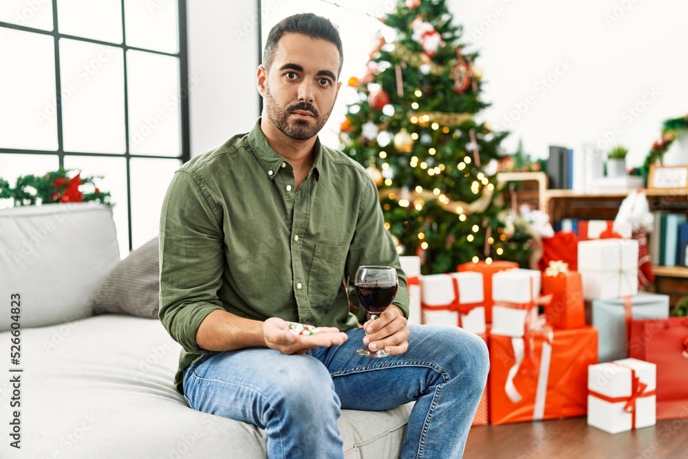 Young hispanic man holding glass of wine and pills sitting on sofa with dog by christmas tree at home