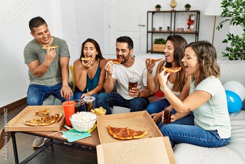 Group of young hispanic friends smiling happy eating italian pizza sitting on the sofa at home.