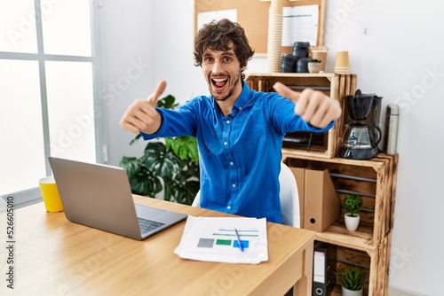 Young hispanic man with beard working at the office using computer laptop approving doing positive gesture with hand, thumbs up smiling and happy for success. winner gesture.