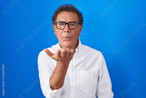 Middle age hispanic man standing over blue background looking at the camera blowing a kiss with hand on air being lovely and sexy. love expression.