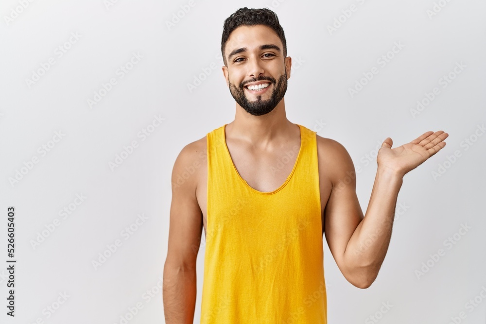 Young handsome man with beard standing over isolated background smiling cheerful presenting and pointing with palm of hand looking at the camera. Young handsome man with beard standing over isolated background smiling cheerful presenting and pointing with palm of hand looking at the camera.