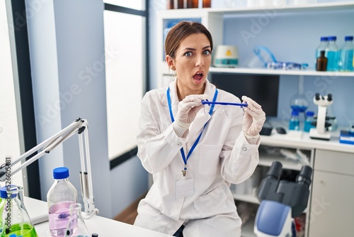 Young brunette woman working at scientist laboratory wearing safety glasses in shock face  looking skeptical and sarcastic  surprised with open mouth