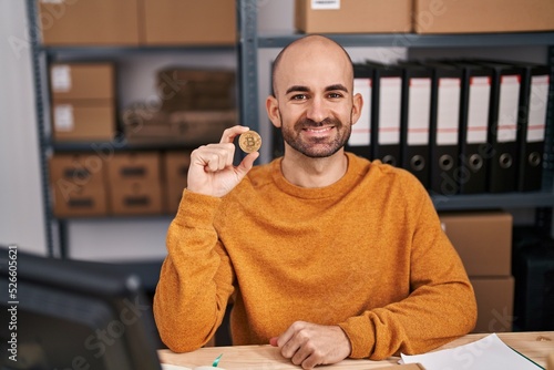 Young bald man with beard working at small business ecommerce holding bitcoin looking positive and happy standing and smiling with a confident smile showing teeth