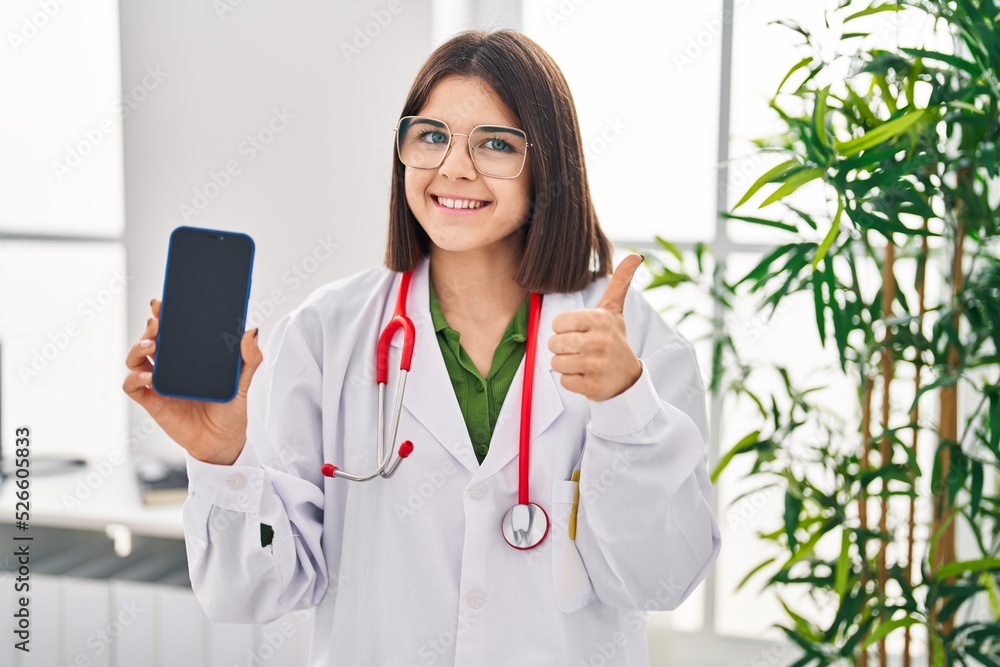 Young hispanic doctor woman showing smartphone screen smiling happy and positive, thumb up doing excellent and approval sign Young hispanic doctor woman showing smartphone screen smiling happy and positive, thumb up doing excellent and approval sign