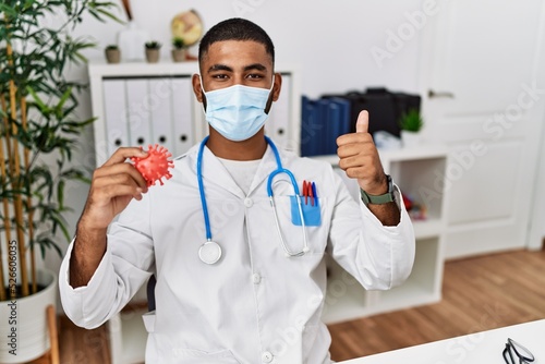Young indian doctor holding virus toy wearing safety mask smiling happy and positive  thumb up doing excellent and approval sign