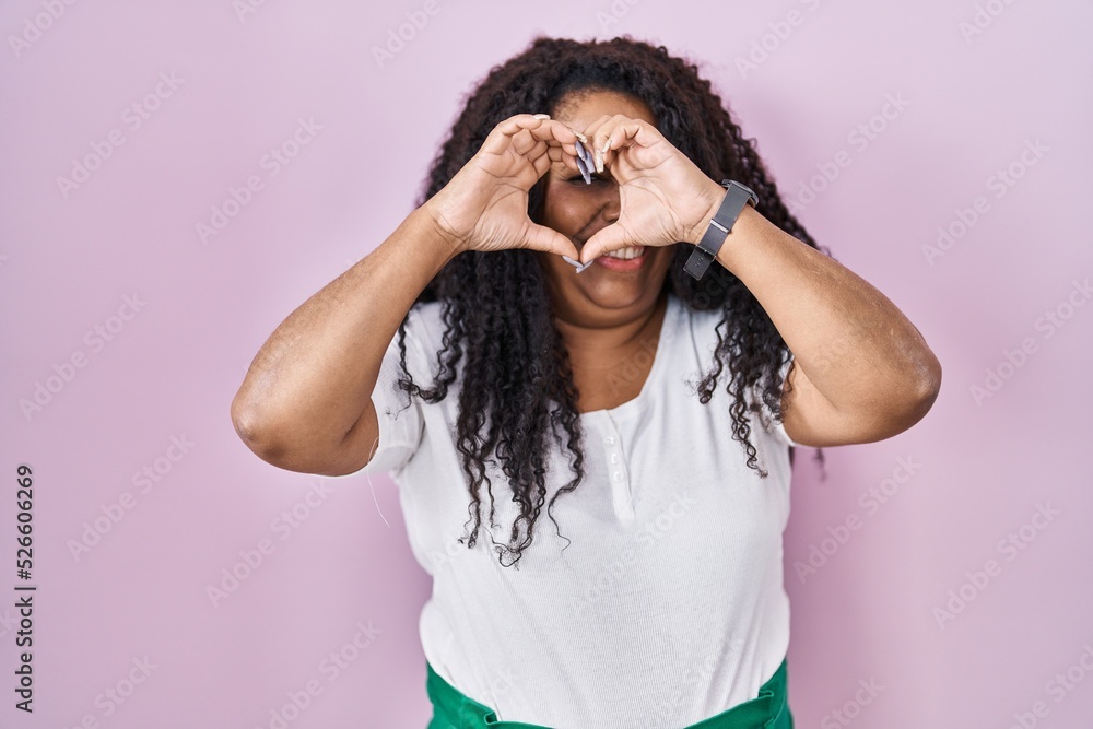 Plus size hispanic woman standing over pink background doing heart shape with hand and fingers smiling looking through sign Plus size hispanic woman standing over pink background doing heart shape with hand and fingers smiling looking through sign