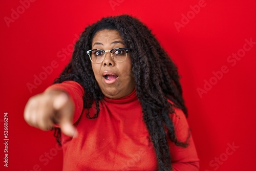 Plus size hispanic woman standing over red background pointing with finger surprised ahead, open mouth amazed expression, something on the front © Krakenimages.com