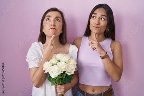 Hispanic mother and daughter holding bouquet of white flowers thinking concentrated about doubt with finger on chin and looking up wondering