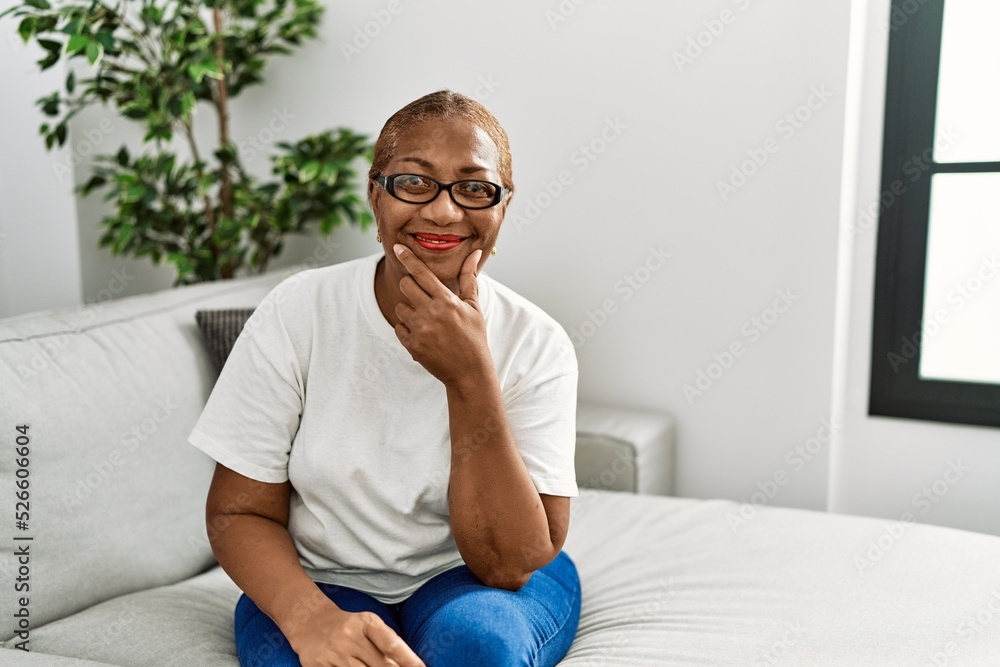 Mature hispanic woman sitting on the sofa at home looking confident at the camera smiling with crossed arms and hand raised on chin. thinking positive. Mature hispanic woman sitting on the sofa at home looking confident at the camera smiling with crossed arms and hand raised on chin. thinking positive.