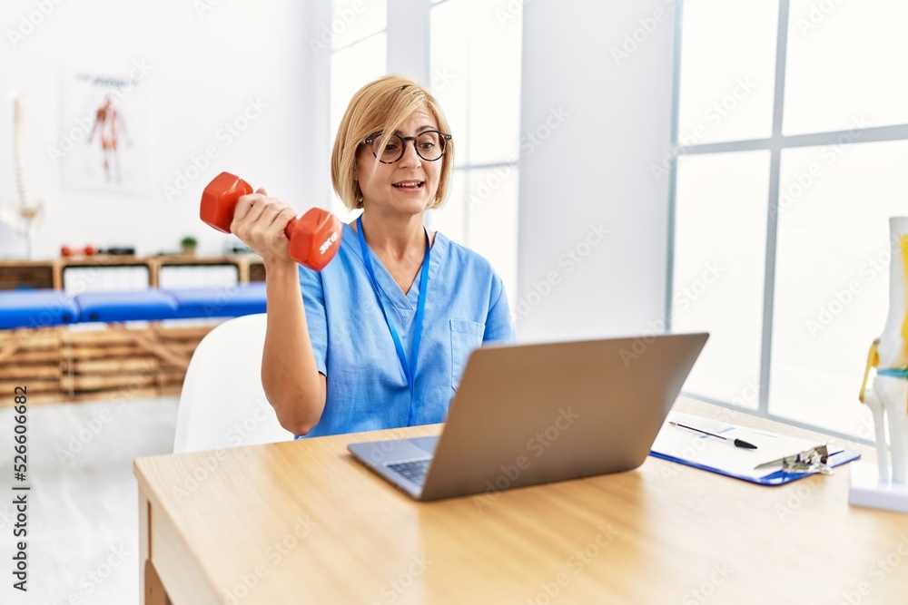 Middle age blonde woman wearing physio therapy uniform doing tele rehab using dumbbells at clinic