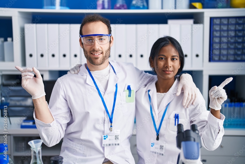 Interracial couple working at scientist laboratory smiling happy pointing with hand and finger to the side Interracial couple working at scientist laboratory smiling happy pointing with hand and finger to the side