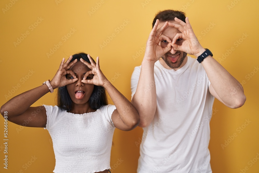 Interracial couple standing over yellow background doing ok gesture like binoculars sticking tongue out, eyes looking through fingers. crazy expression. Interracial couple standing over yellow background doing ok gesture like binoculars sticking tongue out, eyes looking through fingers. crazy expression.