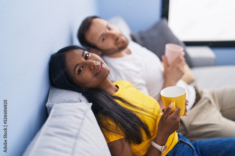 Man and woman interracial couple drinking coffee sitting on sofa at home