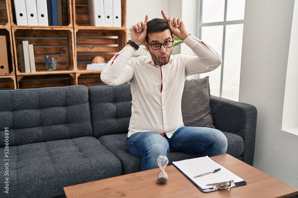 Young hispanic man with beard working at consultation office doing funny gesture with finger over head as bull horns Young hispanic man with beard working at consultation office doing funny gesture with finger over head as bull horns