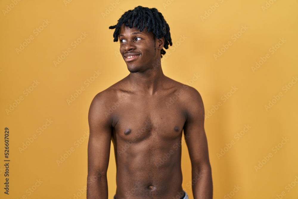 Fototapeta premium Young african man with dreadlocks standing shirtless looking away to side with smile on face, natural expression. laughing confident.