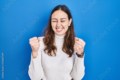 Young hispanic woman standing over blue background excited for success with arms raised and eyes closed celebrating victory smiling. winner concept.