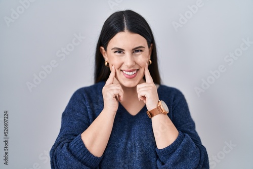 Young brunette woman standing over isolated background smiling with open mouth, fingers pointing and forcing cheerful smile