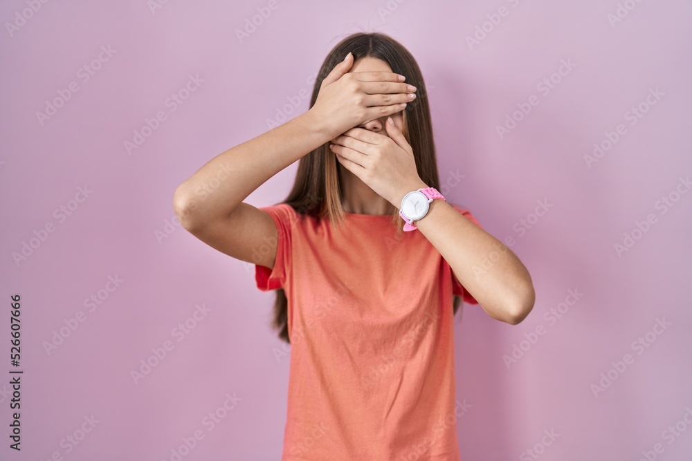 Teenager girl standing over pink background covering eyes and mouth with hands, surprised and shocked. hiding emotion Teenager girl standing over pink background covering eyes and mouth with hands, surprised and shocked. hiding emotion