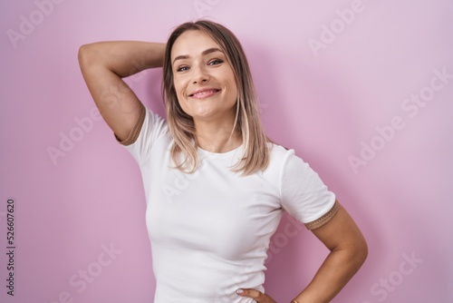 Blonde caucasian woman standing over pink background smiling confident touching hair with hand up gesture  posing attractive and fashionable